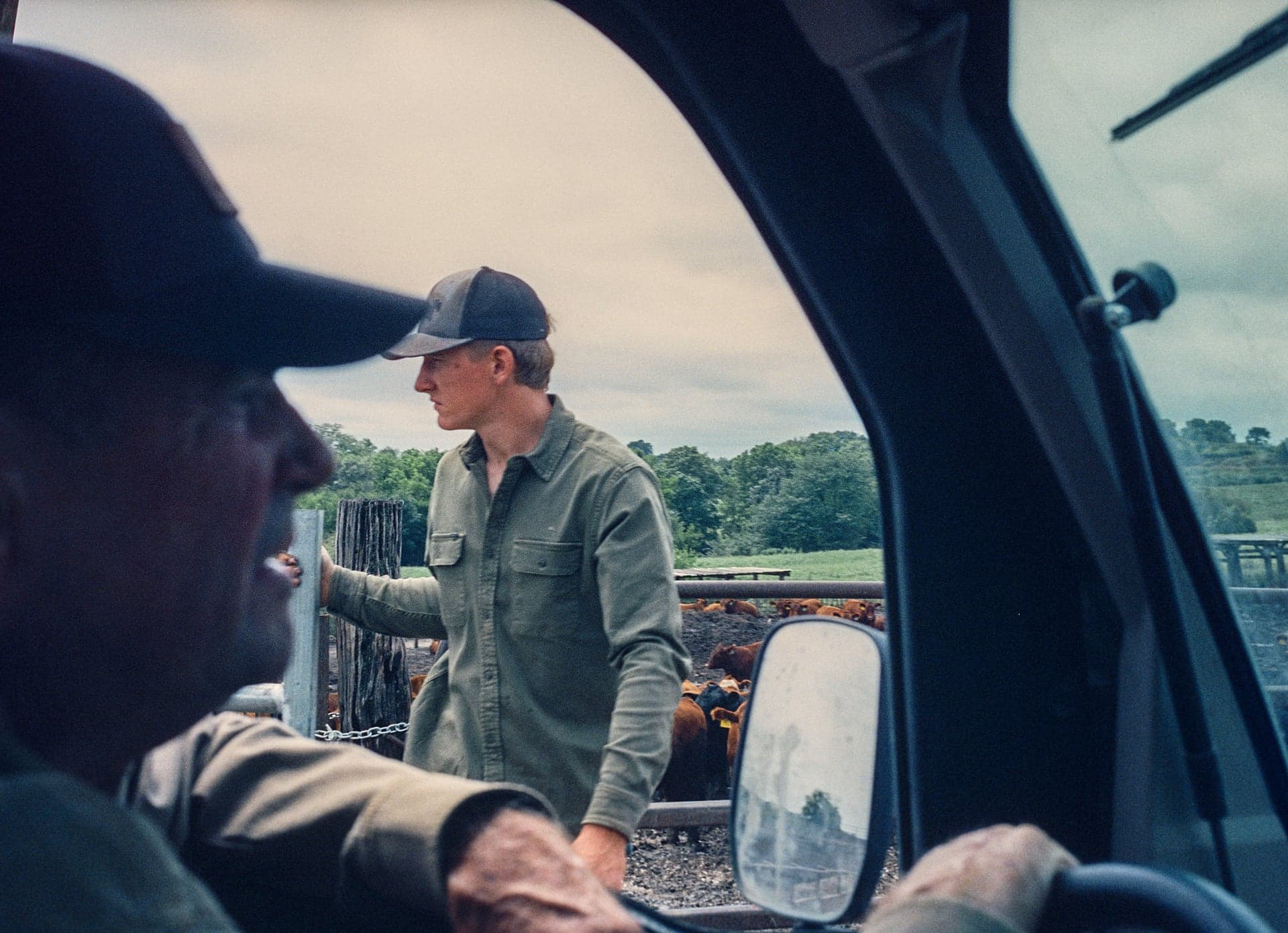 People in a vehicle speak with a man wearing a button-up shirt outdoors beside a fence