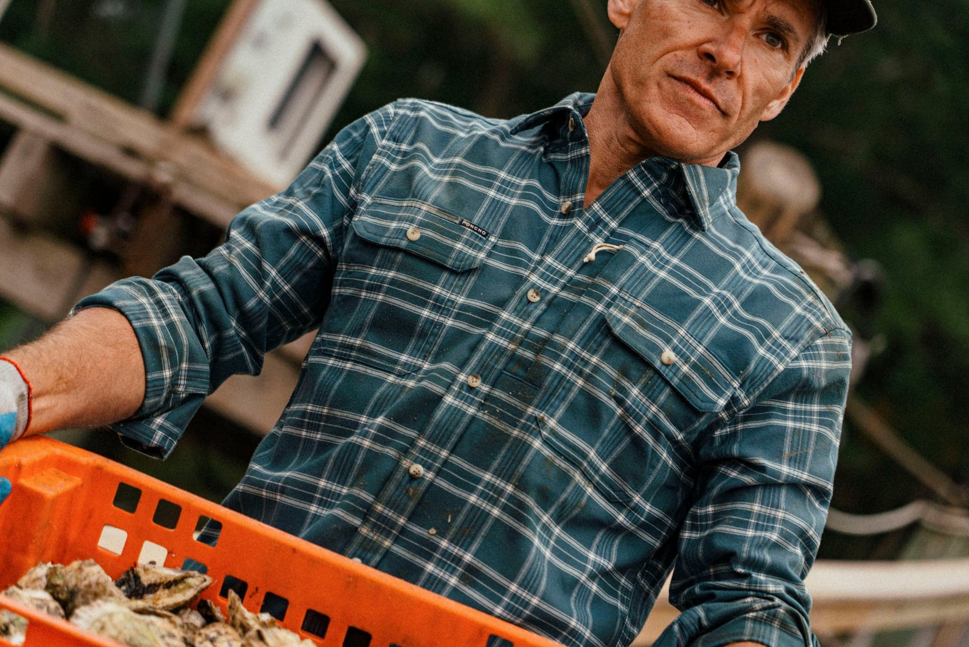 Man wearing a blue plaid outdoor shirt carries an orange crate of shellfish outdoors